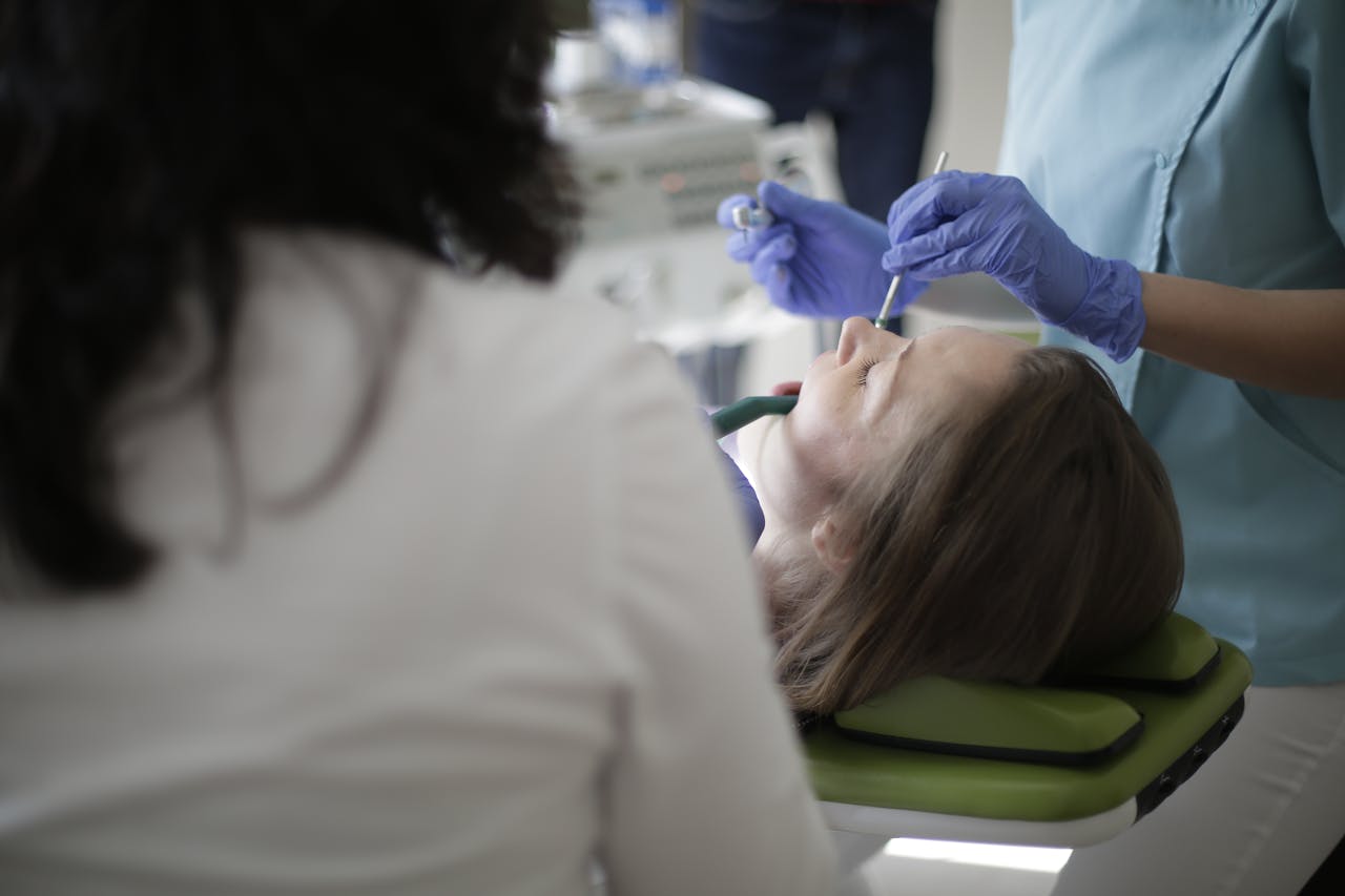 contact-img Crop anonymous female dentist in blue uniform and latex gloves using drill and mouth mirror while treating teeth of female patient lying in dental chair with opened mouth and closed eyes